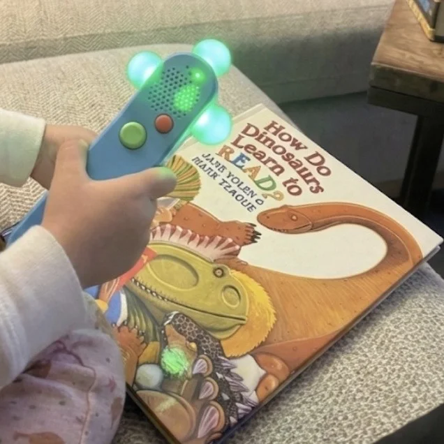 Child's hand holding a blue interactive wand with glowing LEDs, pointing at the How Do Dinosaurs Learn to Read book